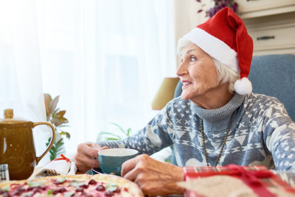 Senior woman in Santa hat at long-term care facility celebrating holidays, drinking tea and looking out the window