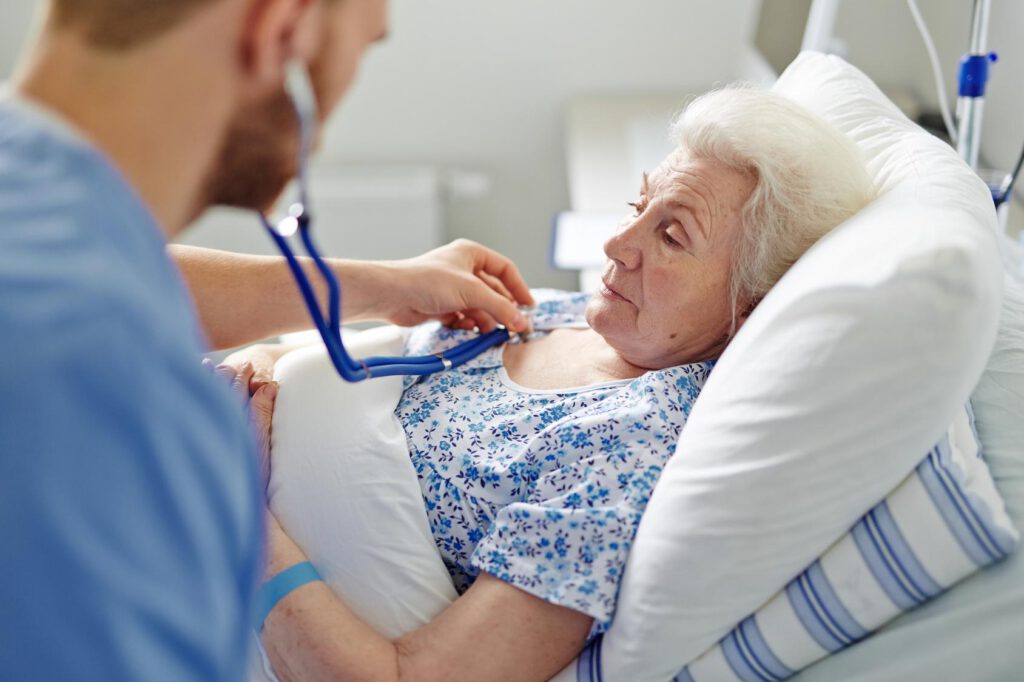 Senior woman lying in hospital bed with doctor listening to heartbeat